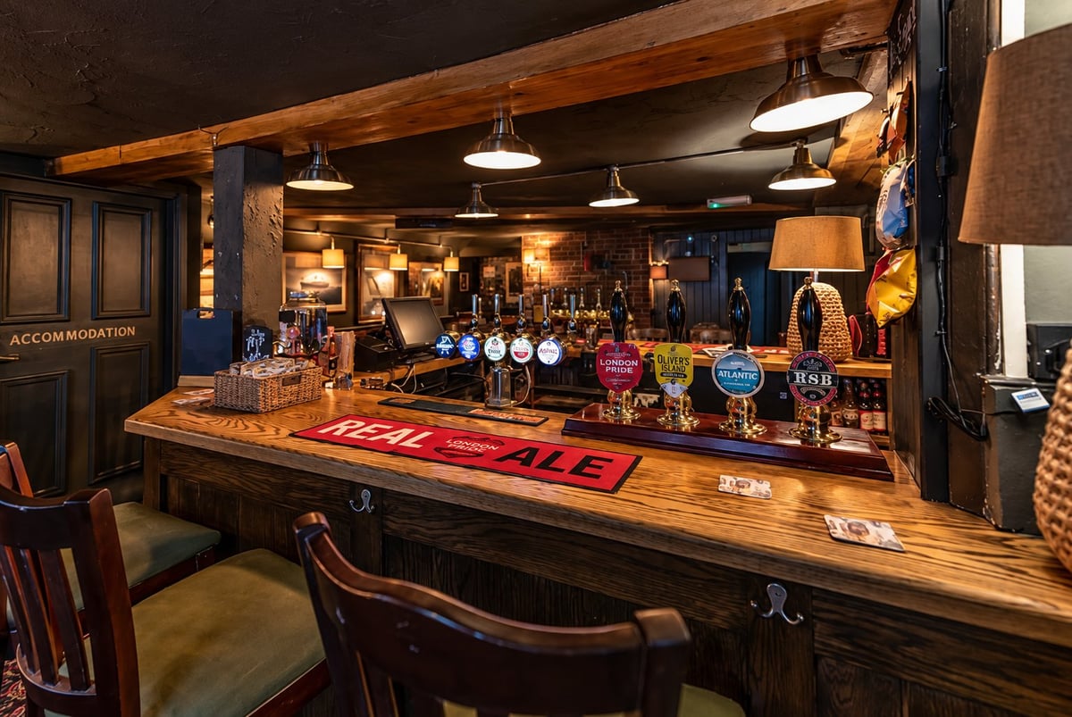 Rustic wooden bar counter with stools and shelves of liquor bottles, pendant lights, and exposed beam ceiling